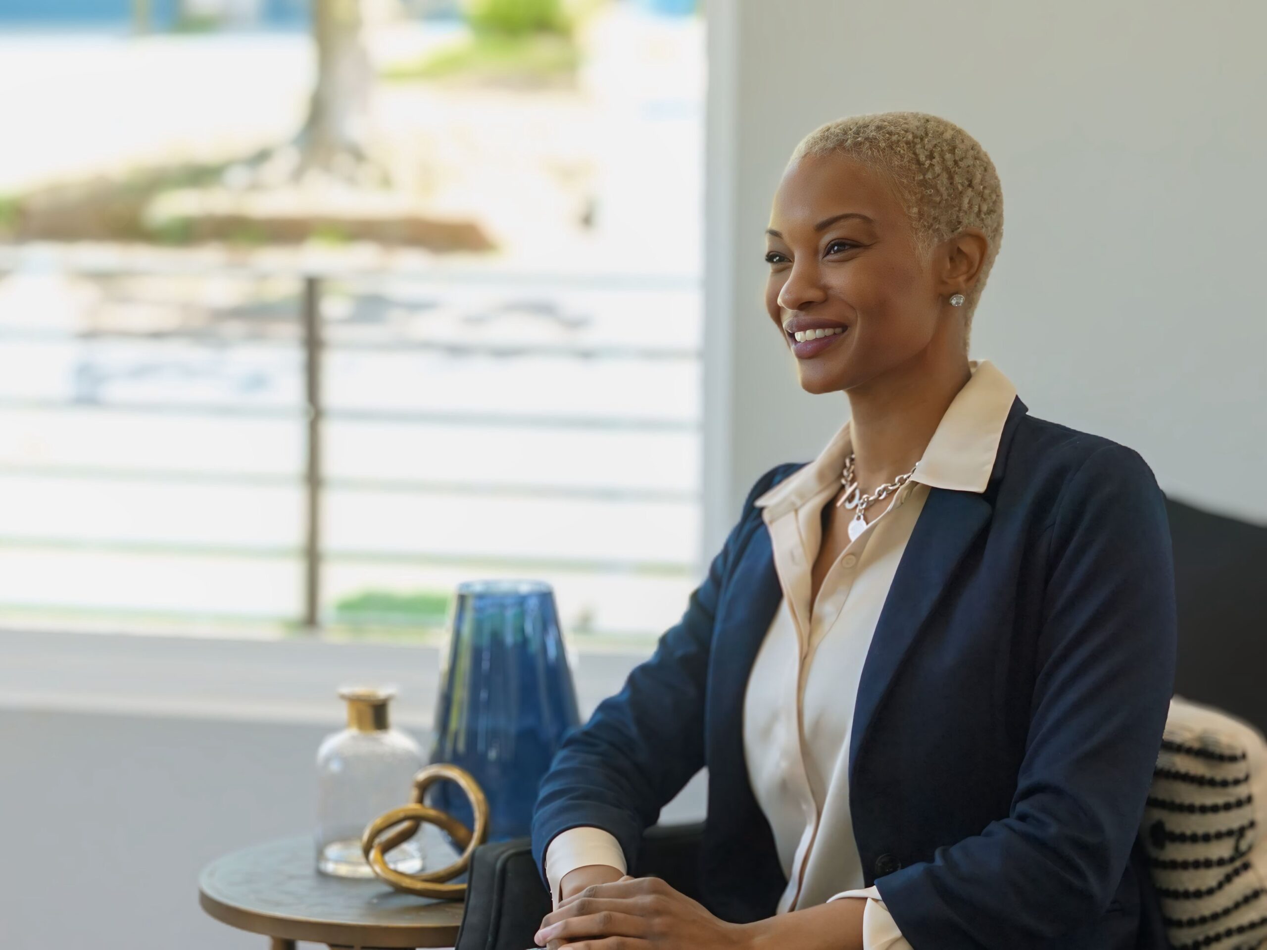 A professional woman with short blonde hair, wearing a navy blazer and cream blouse, smiling confidently while seated at a table with decorative elements in the background. The setting is modern and well-lit, evoking a calm, professional atmosphere.